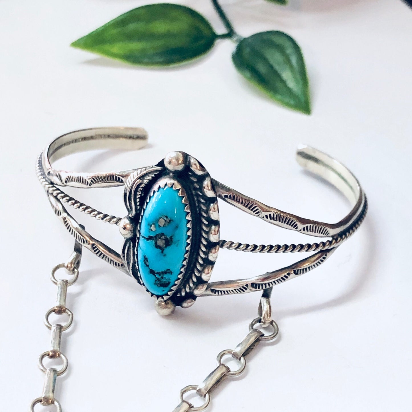 Vintage Navajo silver and turquoise slave bracelet with intricate design and chain detail, set against a white background with green leaf accents, showcasing beautiful bohemian style jewelry.