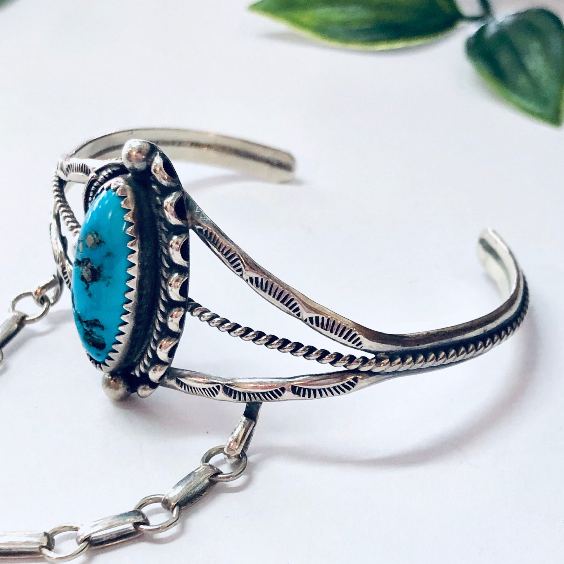 Vintage Navajo silver and turquoise slave bracelet with intricate engraved patterns, a bold blue turquoise stone, and a chain connecting the bracelet to a ring, displayed on a white background with leaves.