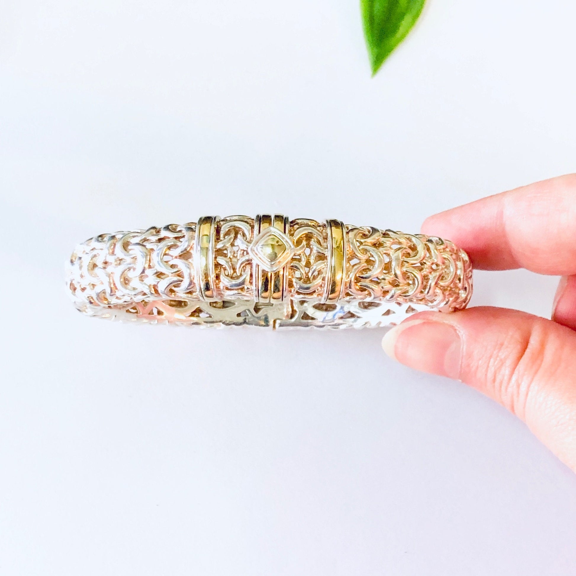 Vintage silver hinged bangle bracelet with gold accents, held in front of a plain background with a green leaf visible at the top of the frame.