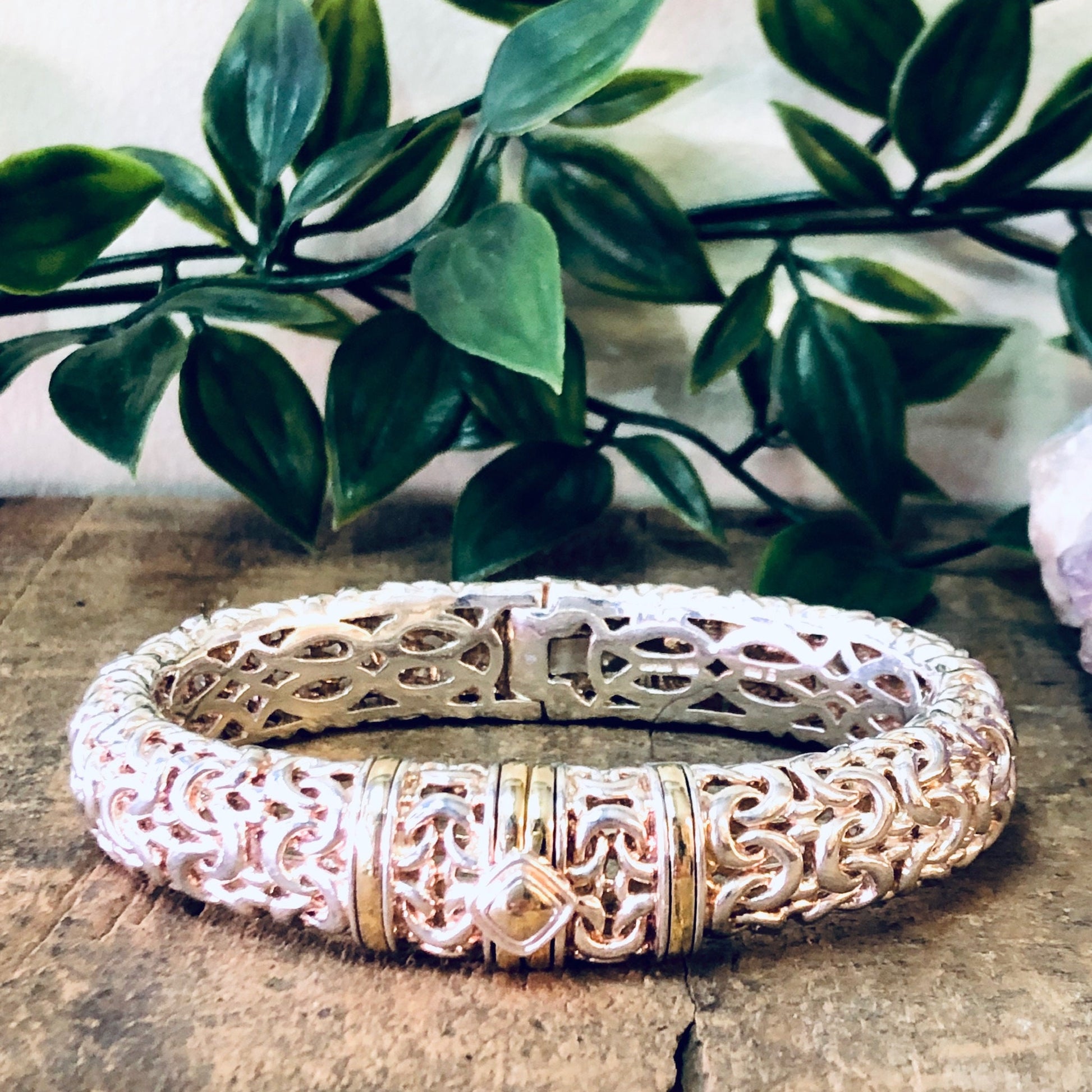 Vintage silver bangle bracelet with intricate cutout design and gold accents, displayed on rustic wooden surface with lush green foliage in the background.