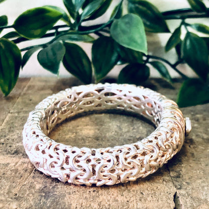 Vintage silver hinged bangle bracelet with intricate filigree design and gold accents placed on stone surface with green foliage in background