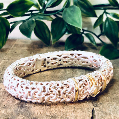 Vintage silver carved bangle bracelet with gold accents and hinged clasp, displayed on stone surface with greenery in background.