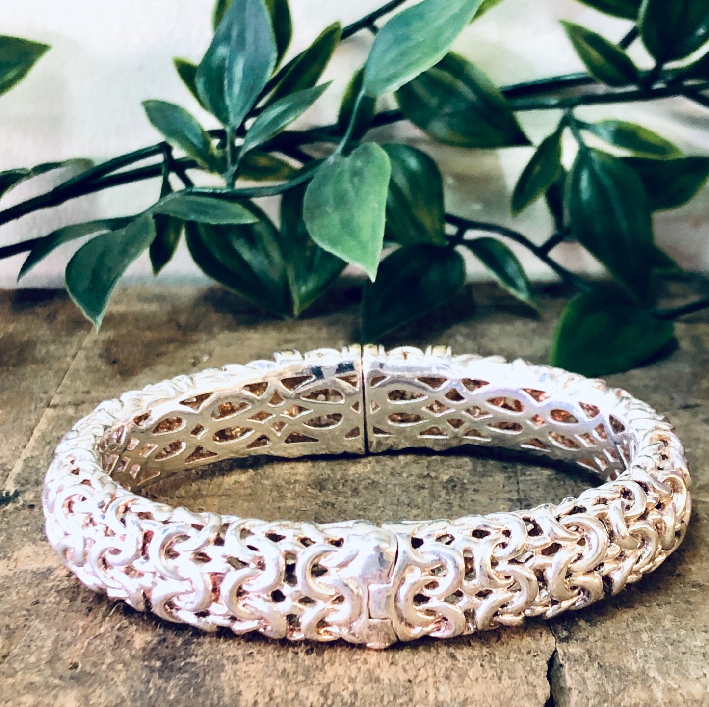 Vintage silver bangle bracelet with intricate cutout design and gold accents, displayed on stone surface with green foliage in background.