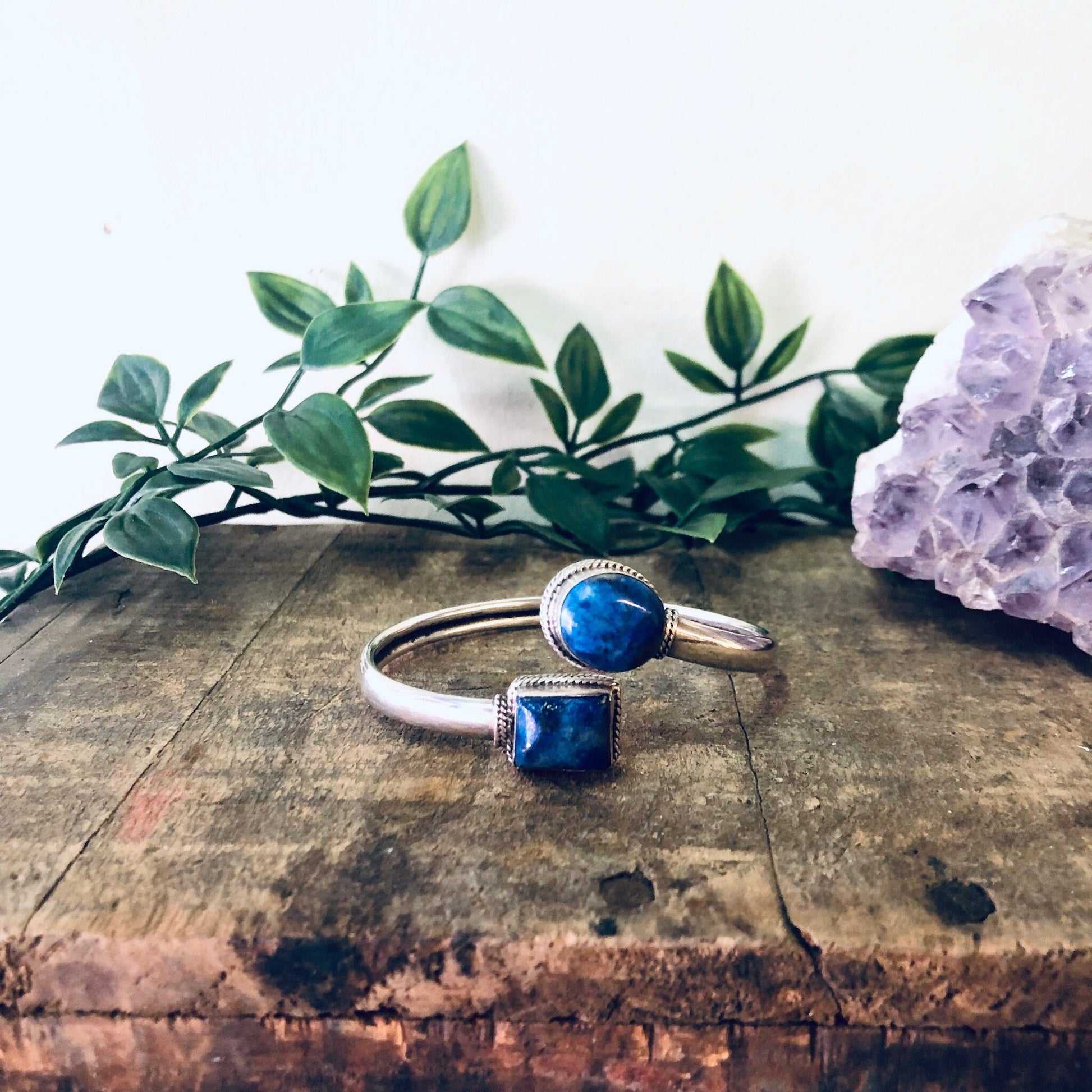 Vintage silver bangle bracelet with blue lapis lazuli gemstones, spiral cuff design, on wooden surface with green leaves and purple crystal rock.