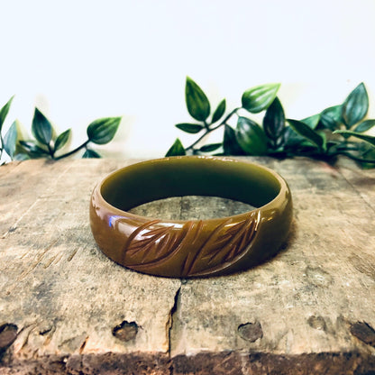 Vintage bakelite bangle bracelet with leaf etching design in shades of green, red and brown, placed on rustic wooden surface with greenery in background.