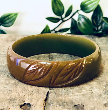 Vintage carved bakelite bangle bracelet in brown with green and red leaf etching design, shown with greenery in background.