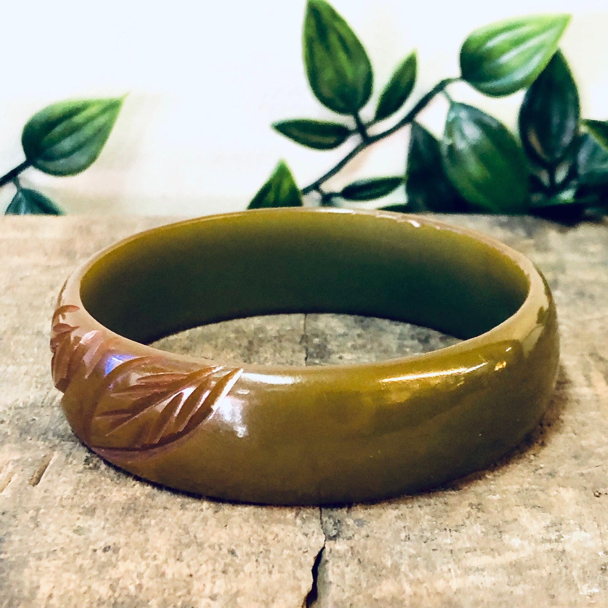 Vintage carved bakelite bangle bracelet in pea soup green with leaf etching detail, on rustic stone background with greenery.