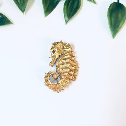 18K yellow gold vintage David Webb seahorse brooch with ruby eye from the 1970s, photographed on a white background with green leaves.