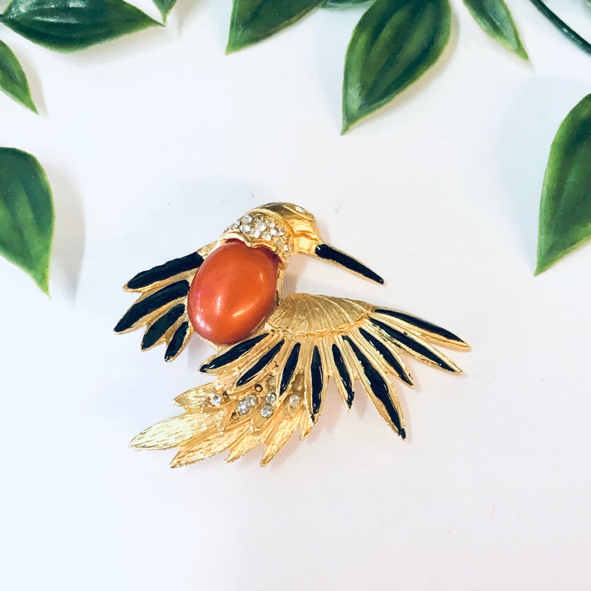 Vintage gold-toned bird brooch with orange stone and black enamel details, surrounded by green leaves on a white background.