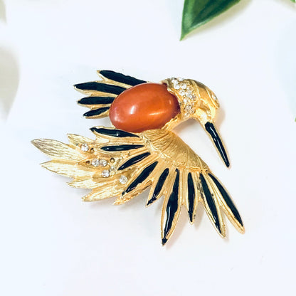 Vintage gold-toned bird brooch pin with black enamel details, orange stone body, and clear rhinestone accents, displayed on a white background with a green leaf.