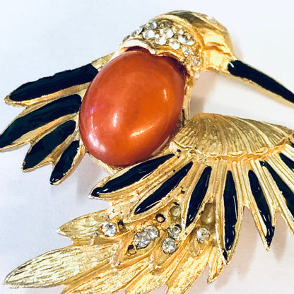 Vintage gold-toned bird brooch pin with orange stone body, black enamel wings, and clear rhinestone accents, depicting a stylized bird in flight against a white background.