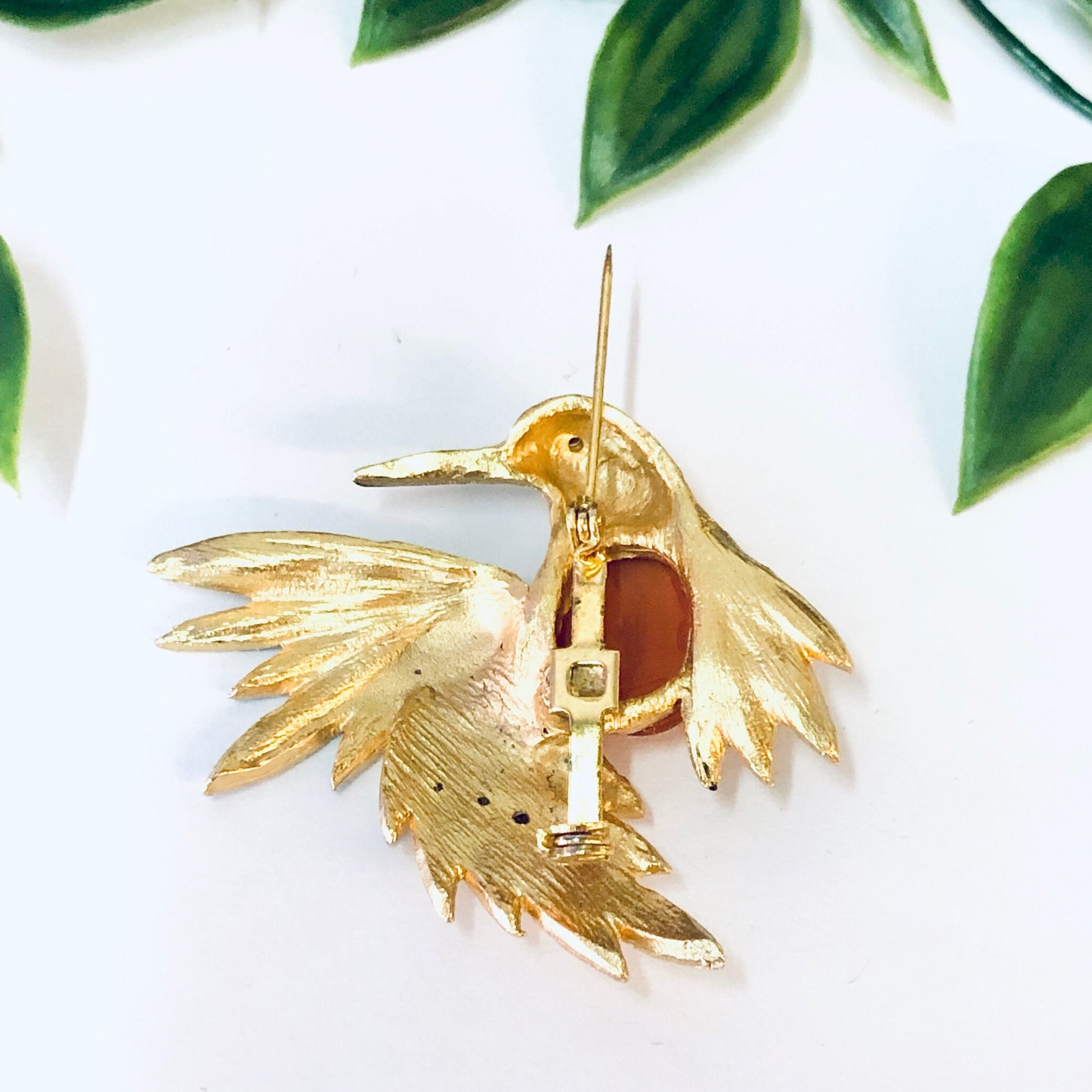 Vintage gold-toned bird brooch pin with orange stone and rhinestones, perched on branch with green leaves in white background.