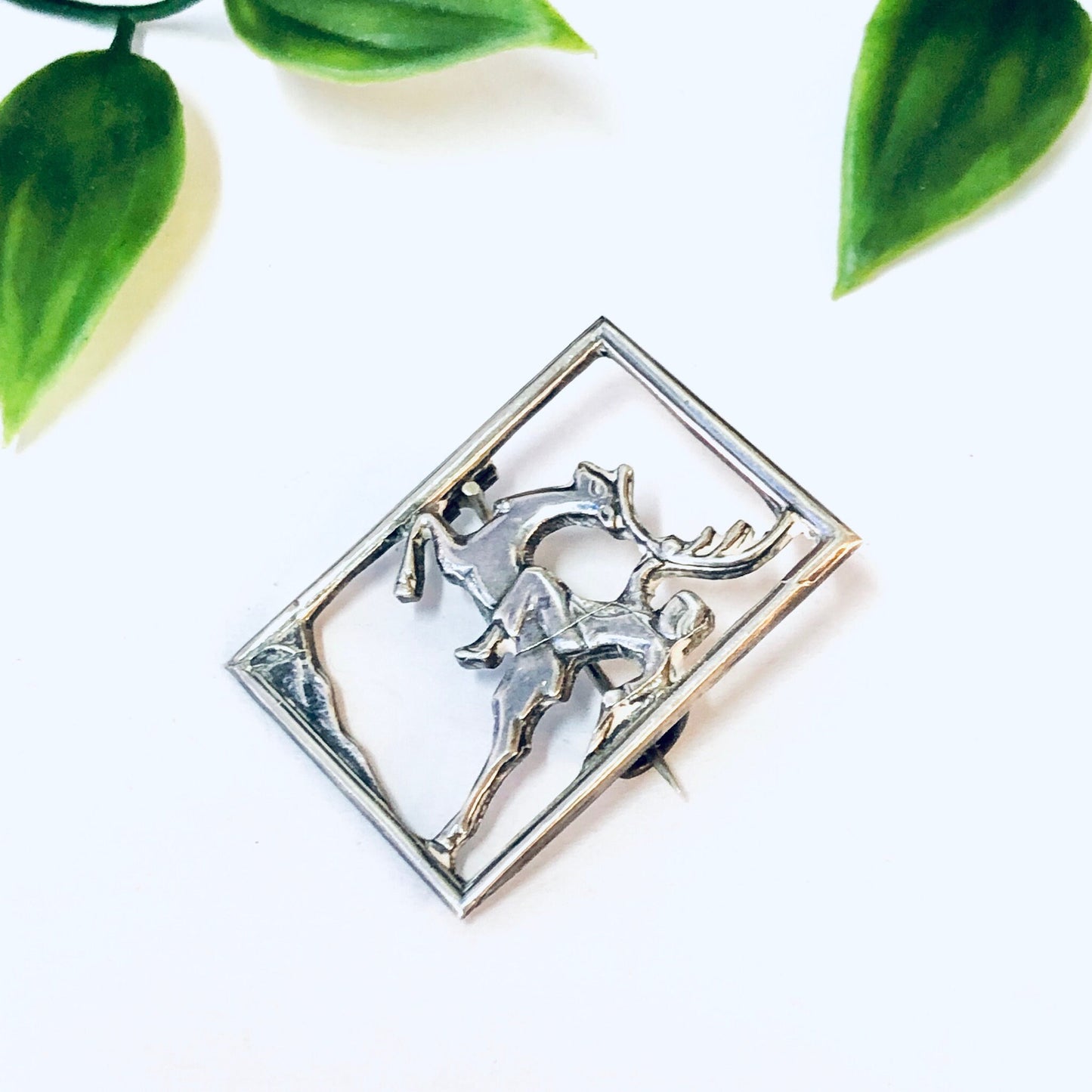 Vintage silver deer brooch featuring a cut-out design with a man riding the deer, surrounded by green leaves on a white background.