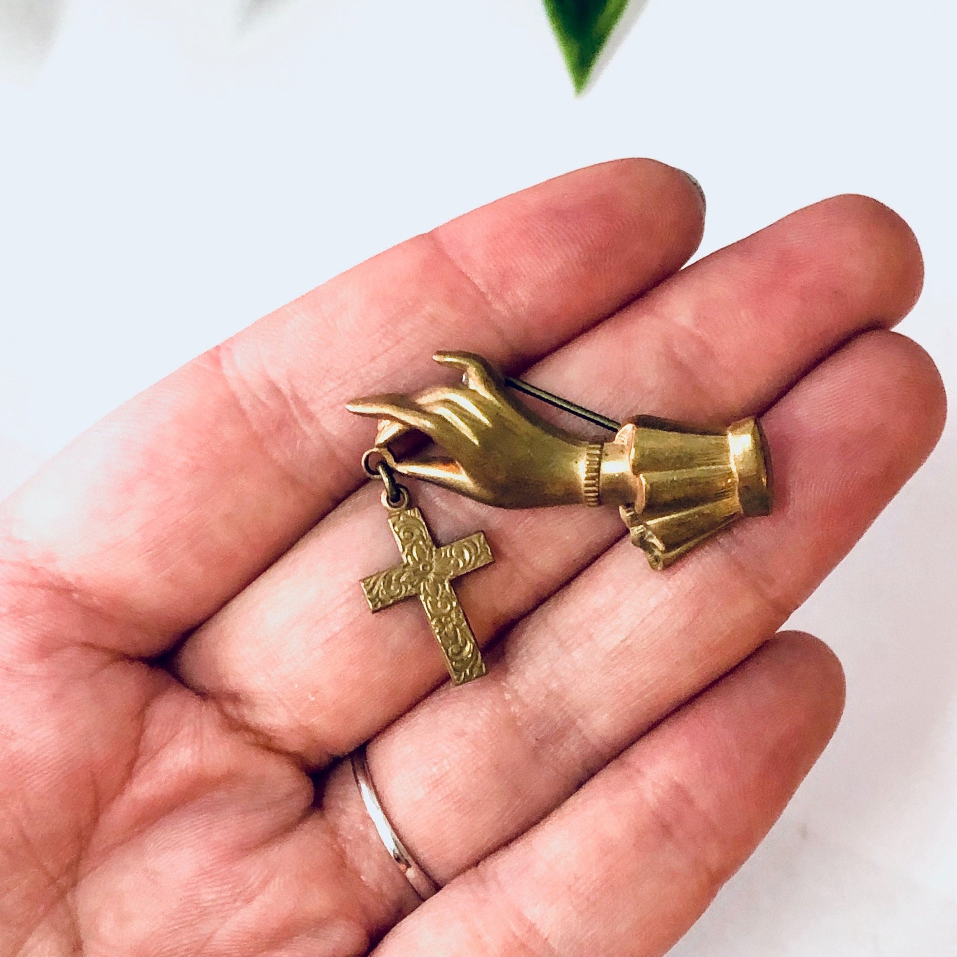 Vintage gold-toned cross brooch shaped like hands clasped together, held in an open human palm against a white background with a green leaf visible at the top of the frame.