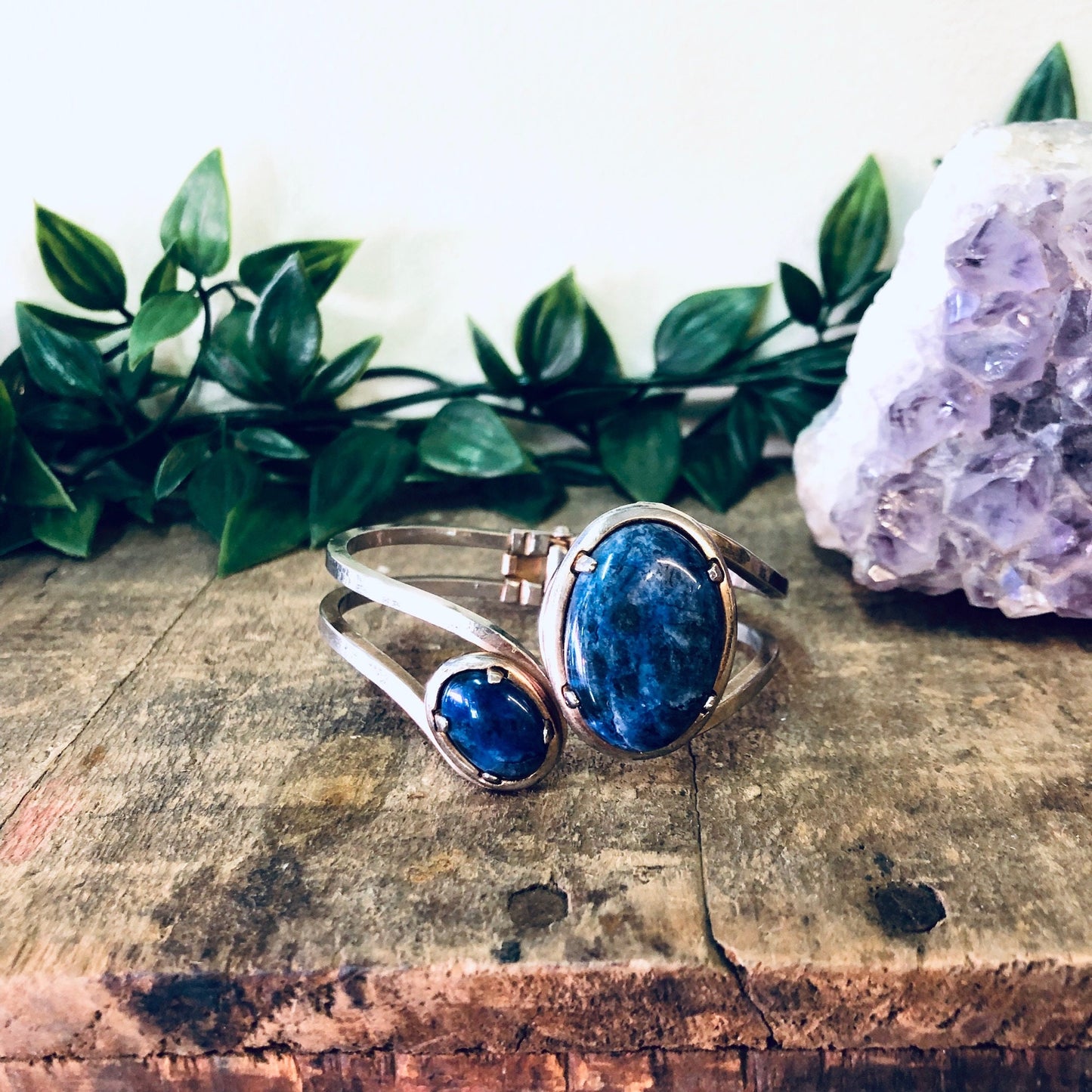 Vintage silver-toned cuff bracelet with blue stones on wooden surface next to amethyst crystal and green leaves