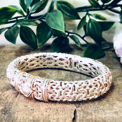 Vintage silver hinged bangle bracelet with intricate cutout designs and gold accents, placed on rustic stone surface with green plant leaves in background.