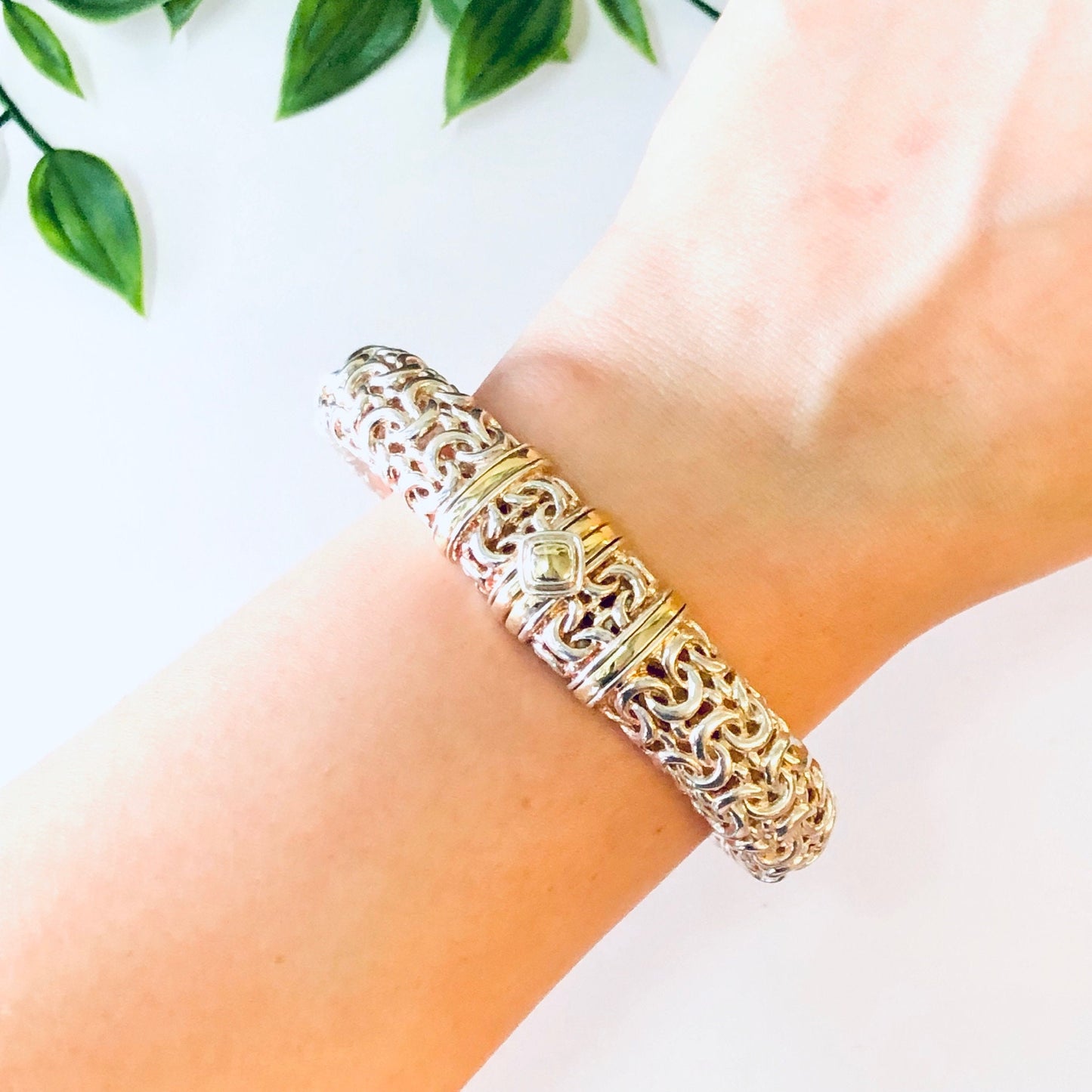 Ornate vintage silver hinged bangle bracelet with intricate gold accent design worn on a wrist, with green leaves in the background.