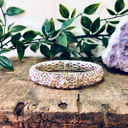 Vintage silver cuff bracelet with intricate gold accents on a rustic wooden surface surrounded by green foliage and amethyst crystal.