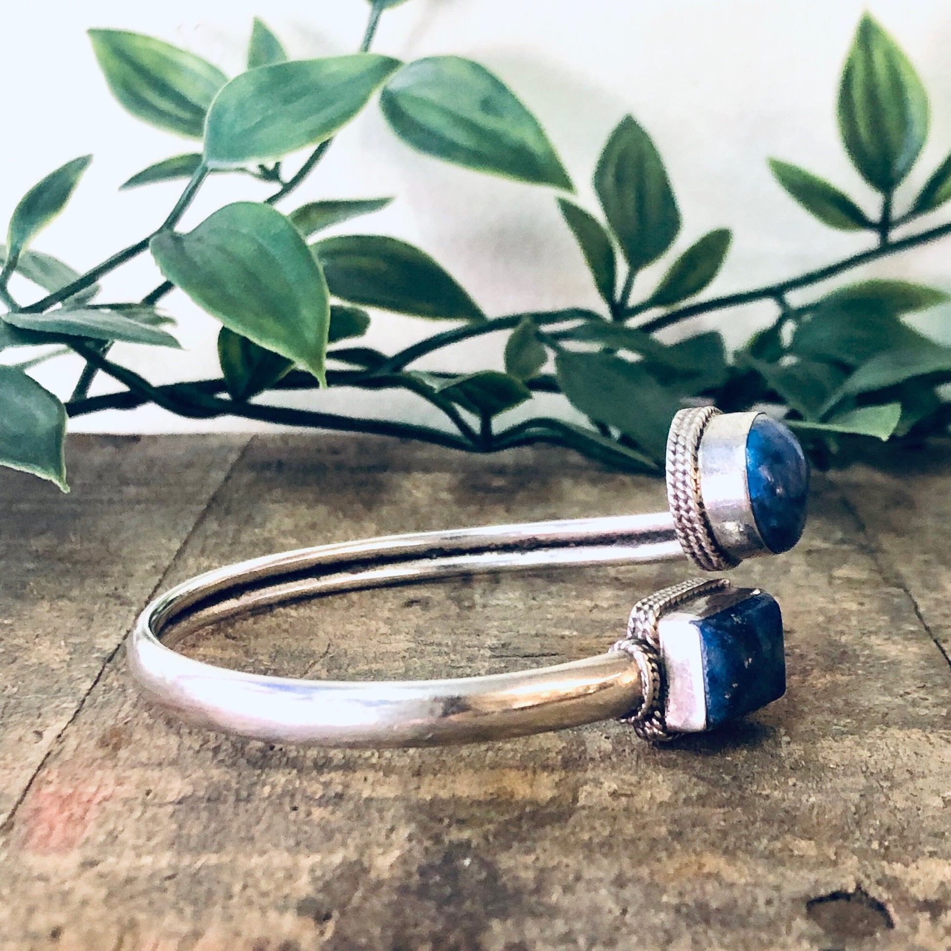 Vintage silver bangle bracelet with two blue lapis lazuli stones, sitting on a wooden surface with green leaves in the background.