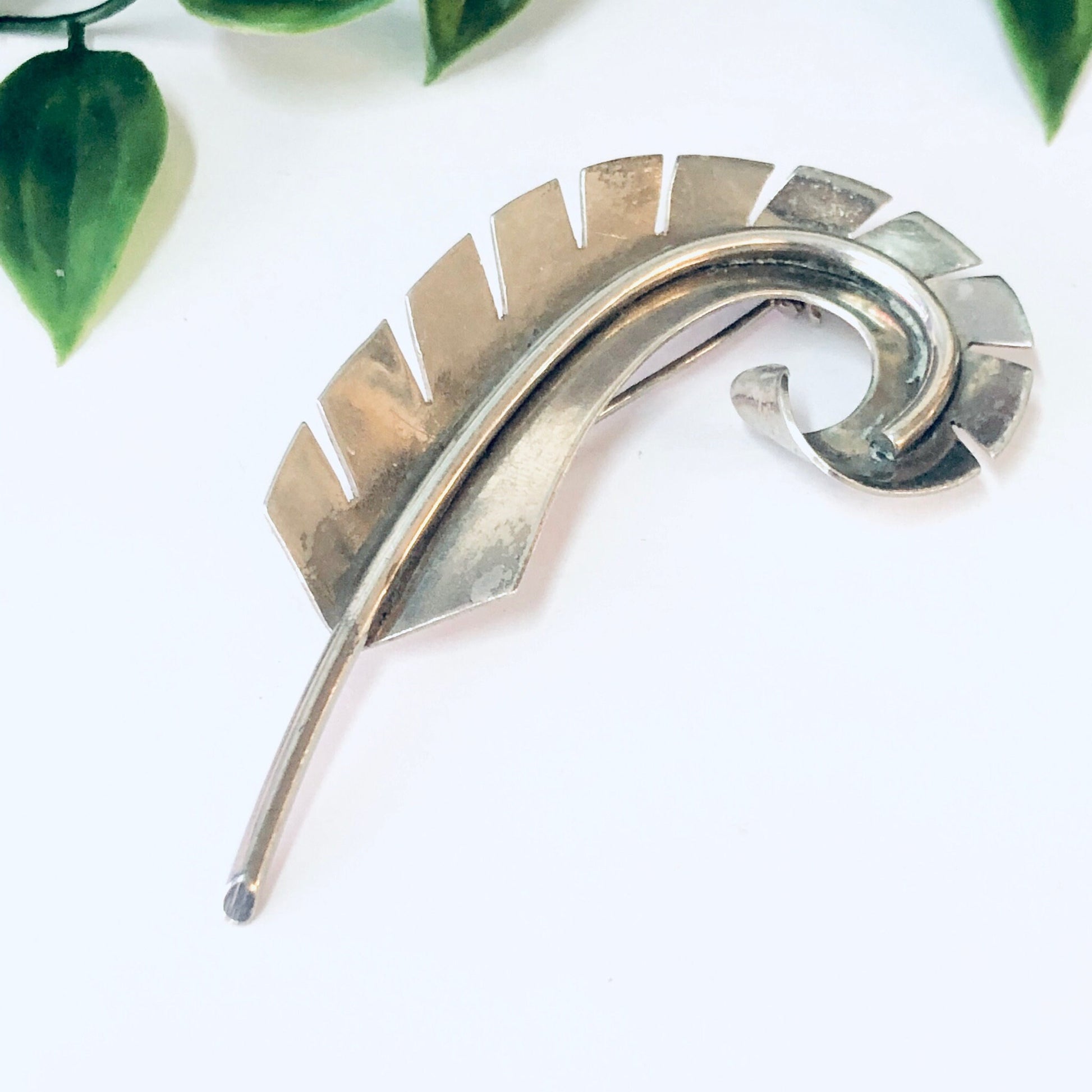 Vintage silver curved feather brooch pin, abstract statement jewelry piece with textured metallic finish, displayed on white background with green foliage.