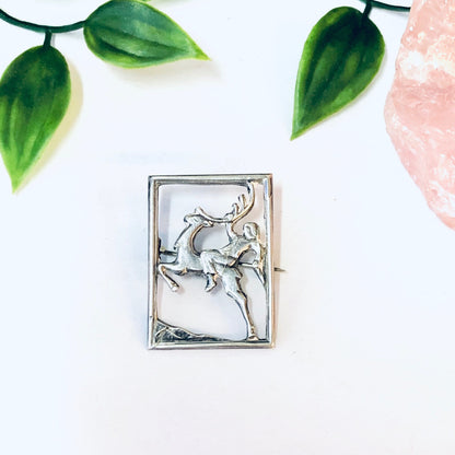 Vintage silver deer brooch featuring a silhouette of a man riding a deer, surrounded by green leaves on a white background.