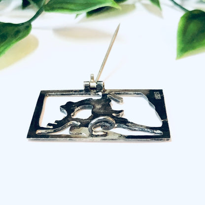 Vintage silver cut-out brooch pin featuring a deer with a person riding on top, displayed on a white surface with green leaf accents in the background.