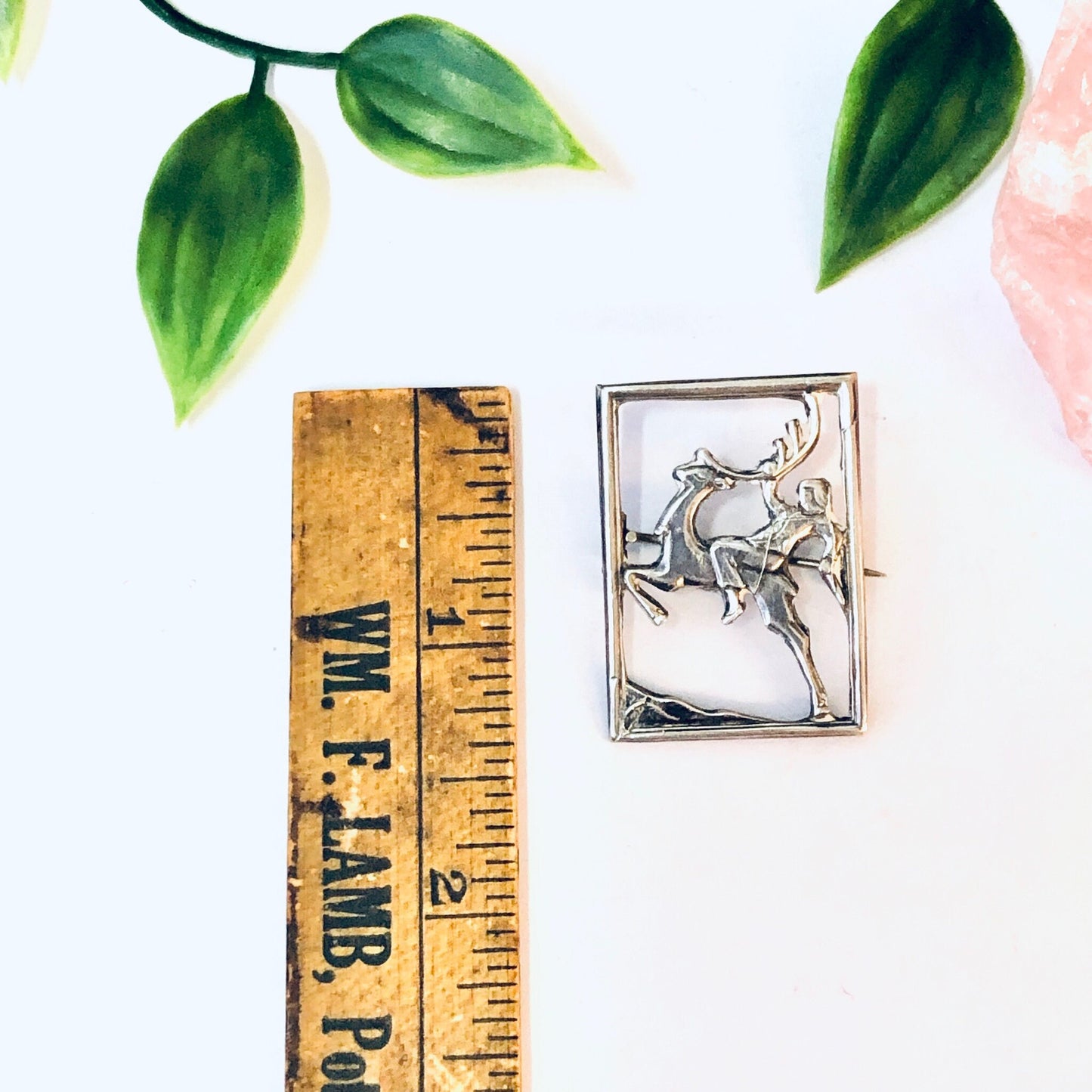 Vintage silver deer brooch with cut-out design featuring a man riding the deer, displayed with a wooden ruler and green leaves on a white background.