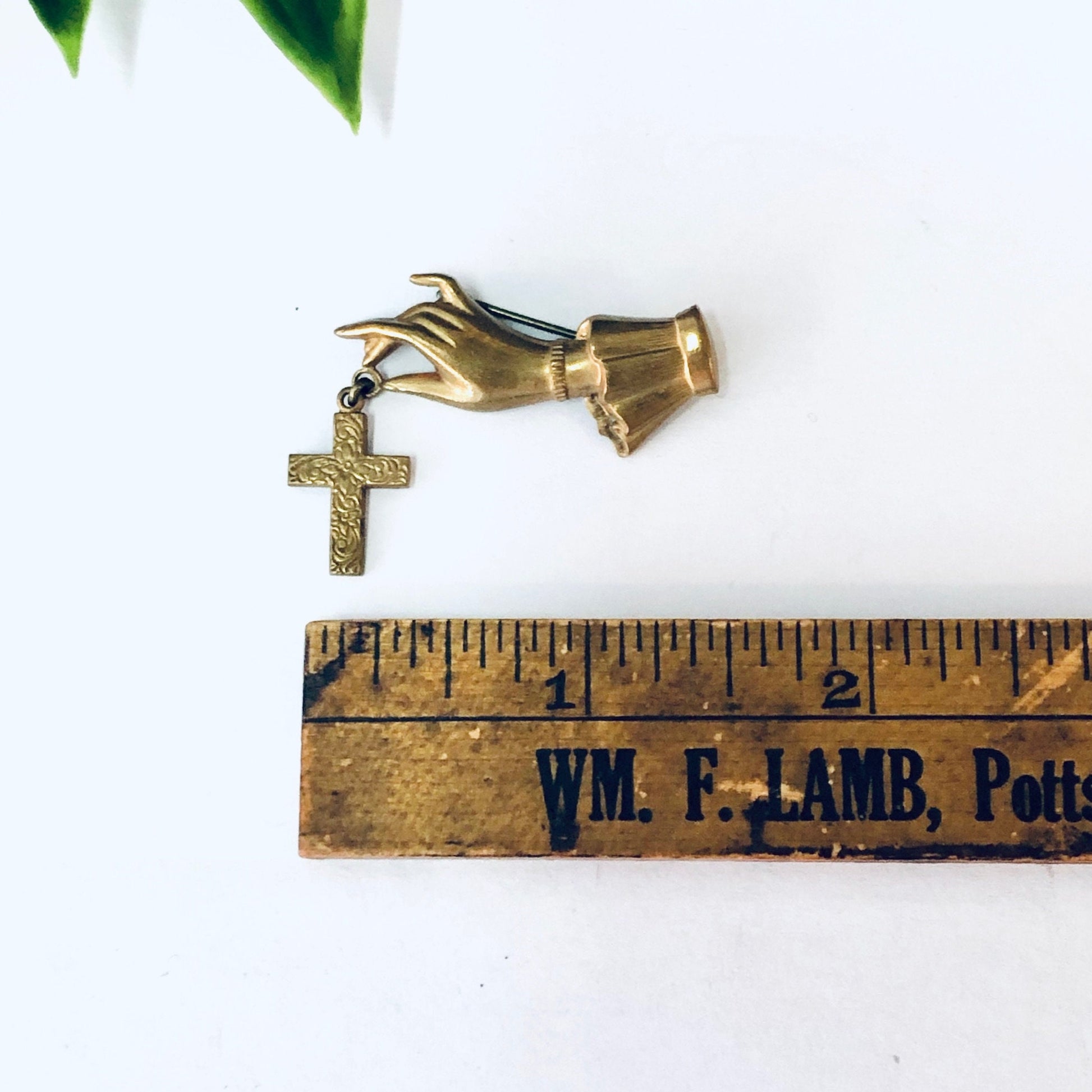 Vintage gold-toned cross brooch shaped like hand holding cross, next to antique wooden ruler with "WM. F. LAMB" text, on white background with leaves.