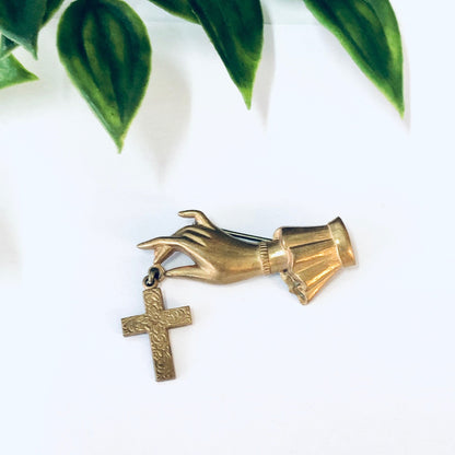 Vintage gold-toned cross and hand brooch pin, religious costume jewelry, displayed on white background with green leaves.