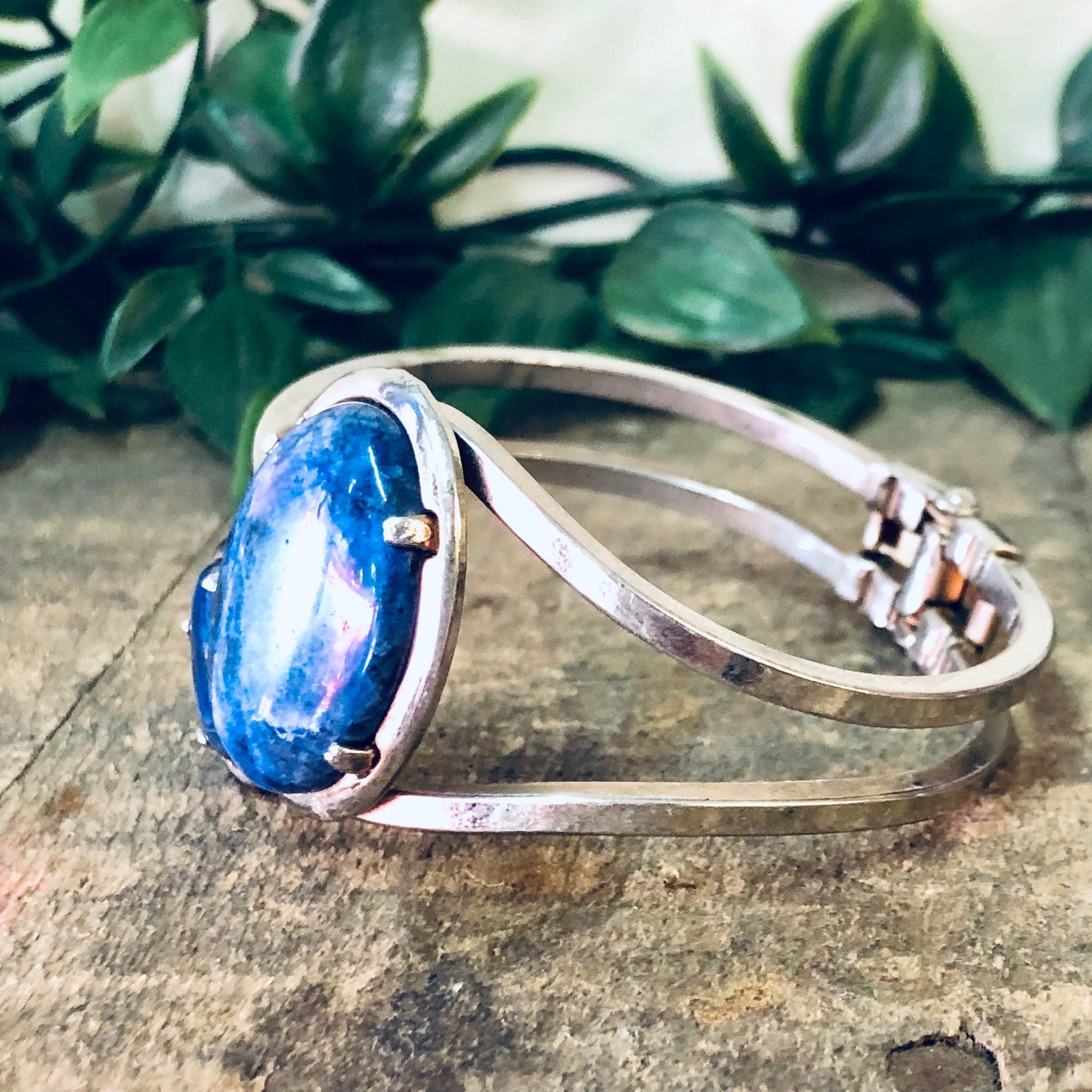 Vintage silver-toned hinged cuff bracelet with large oval blue stone, resting on weathered wood surface with green foliage in background.