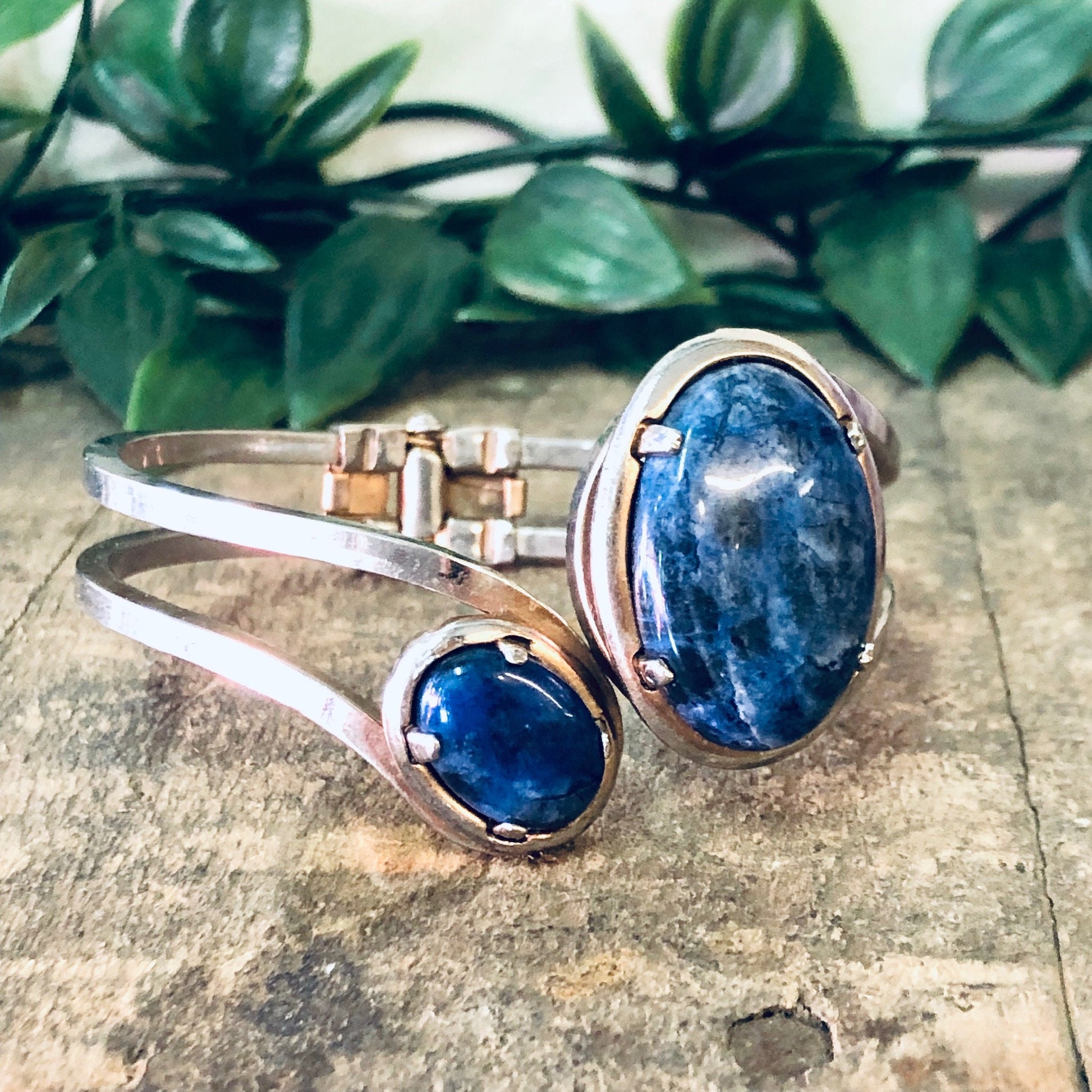 Vintage silver-toned hinged cuff bracelet with two oval blue stones, displayed on a wooden surface with greenery in the background.