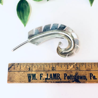 Vintage silver abstract curved feather pin brooch jewelry from Wm. F. Lamb, Pottstown, Pa., with ruler for scale against white background with green leaves.