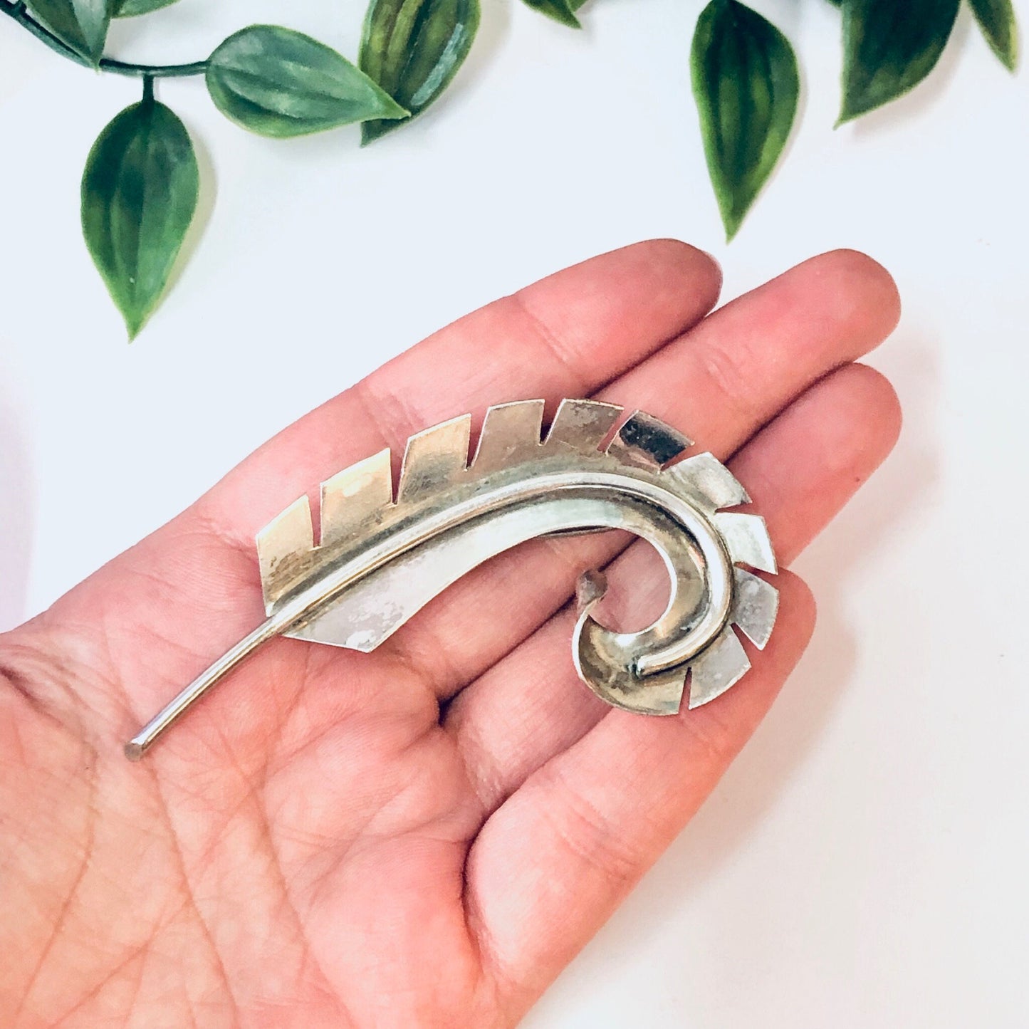 Vintage silver curved feather brooch pin held in palm of hand with green leaves in background