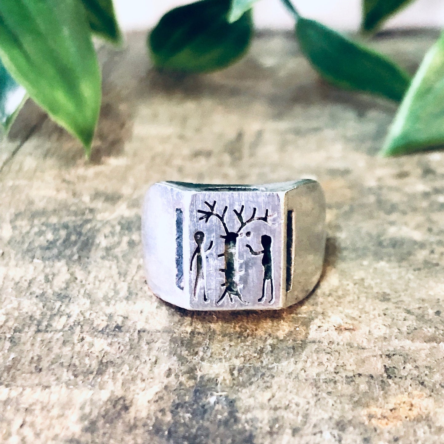 Silver ring with engraved figures resembling Adam, Eve and the Tree of Life from the biblical creation story, placed on a stone surface with green foliage in the background.