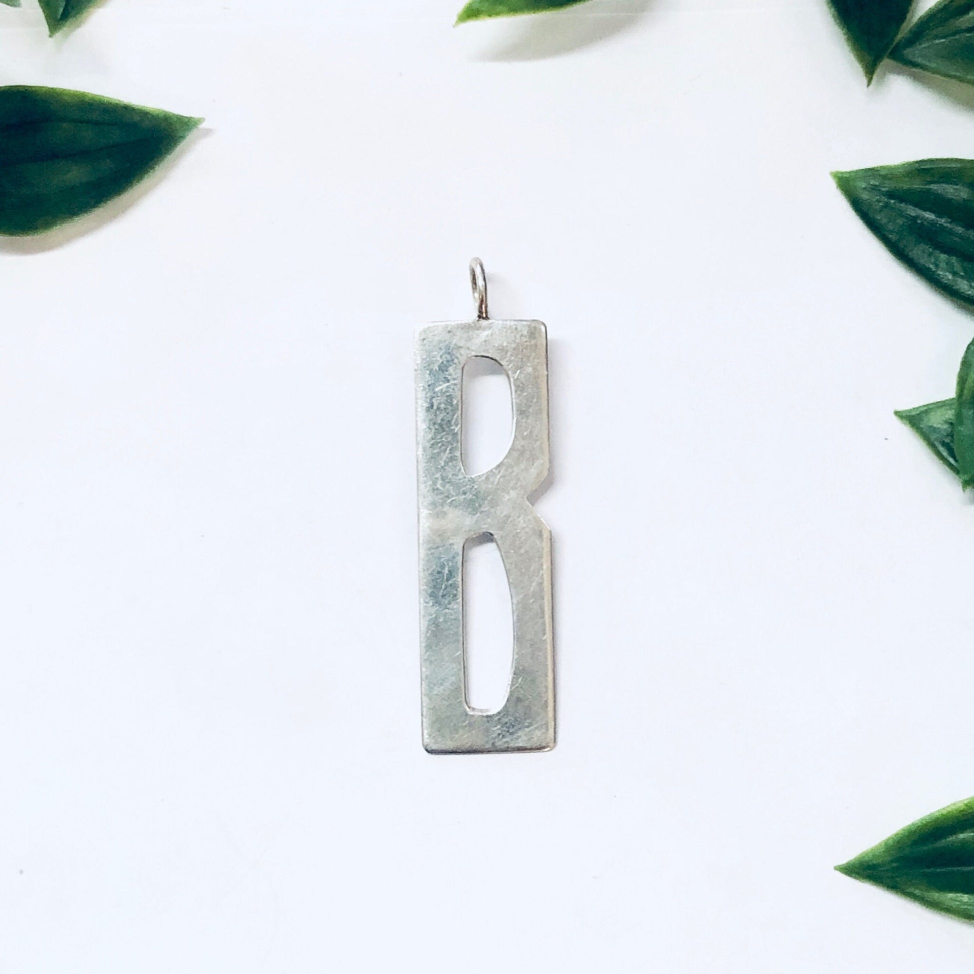 Vintage silver initial "B" pendant by Leonore Doskow, surrounded by green leaves on a white background.