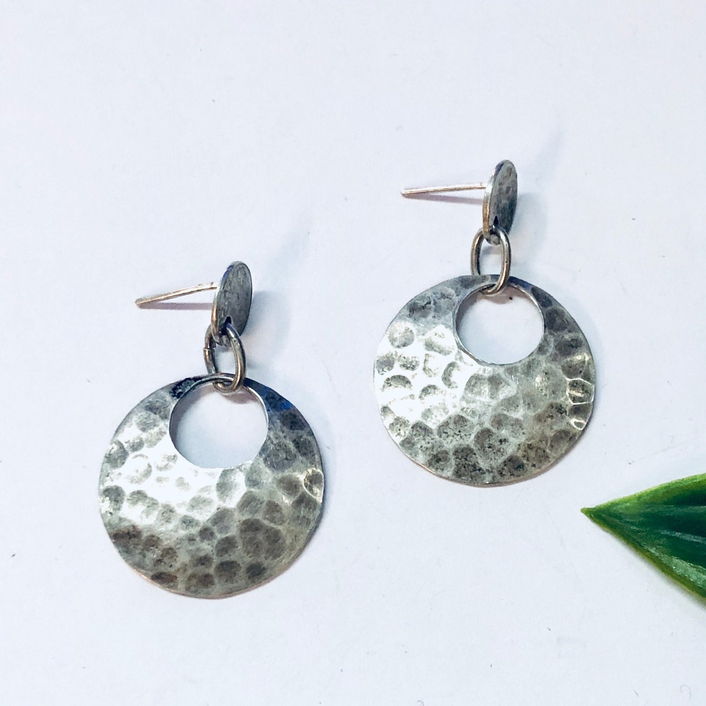 Vintage hammered silver dangle earrings with circular design and pierced ear wires on white background with green leaf.