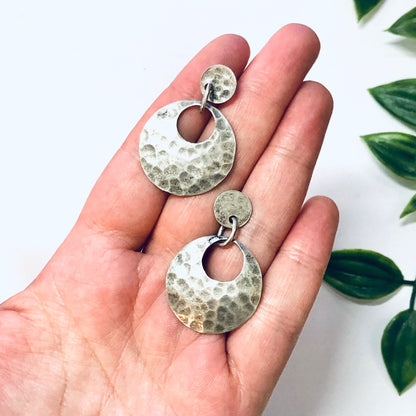 Vintage hammered silver dangle earrings with a unique circular design, held in a person's hand against a white background with green leaves.