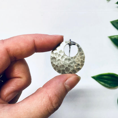 Hammered silver circular vintage dangle earrings held in fingers against white background with green leaves.