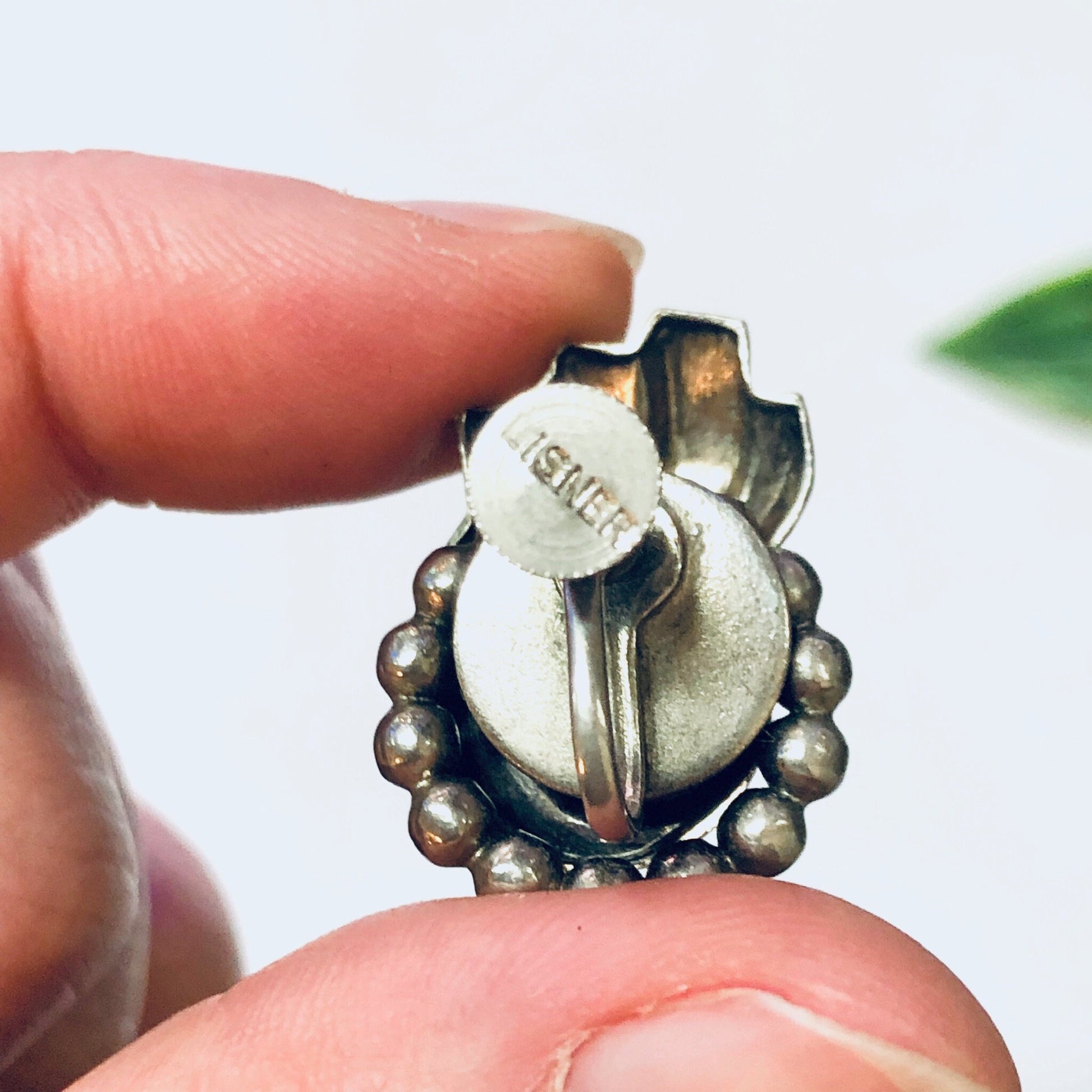 Vintage silver screw back earrings with unique floral design, featuring round beaded detailing around a central circular element engraved with "Lisner", held in a person's fingers against a plain background.