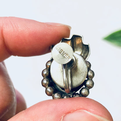 Vintage silver screw back earrings with unique floral design, featuring round beaded detailing around a central circular element engraved with "Lisner", held in a person's fingers against a plain background.