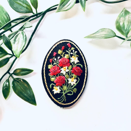Oval vintage crewel brooch with red roses and daisy floral design against a white background with green leaves.