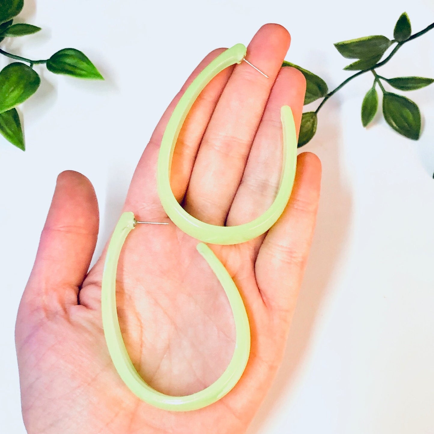 Pair of large neon green vintage dangle earrings from the 1980s held in an open hand, with green leaves in the background.