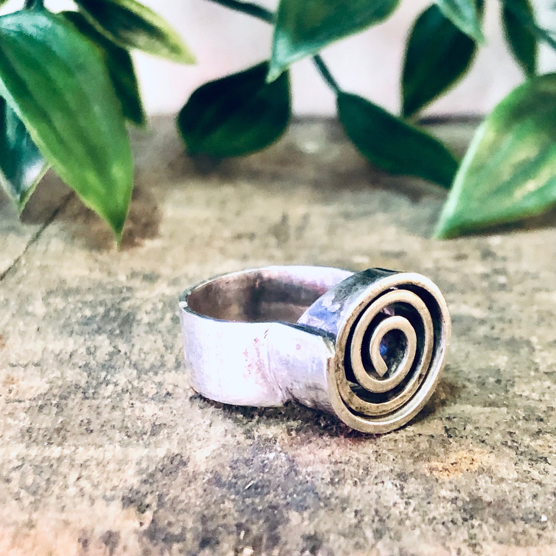 Vintage silver ring with swirl design on a thick band, placed on a textured surface with green leaves in the background, showcasing modernist bohemian jewelry style.