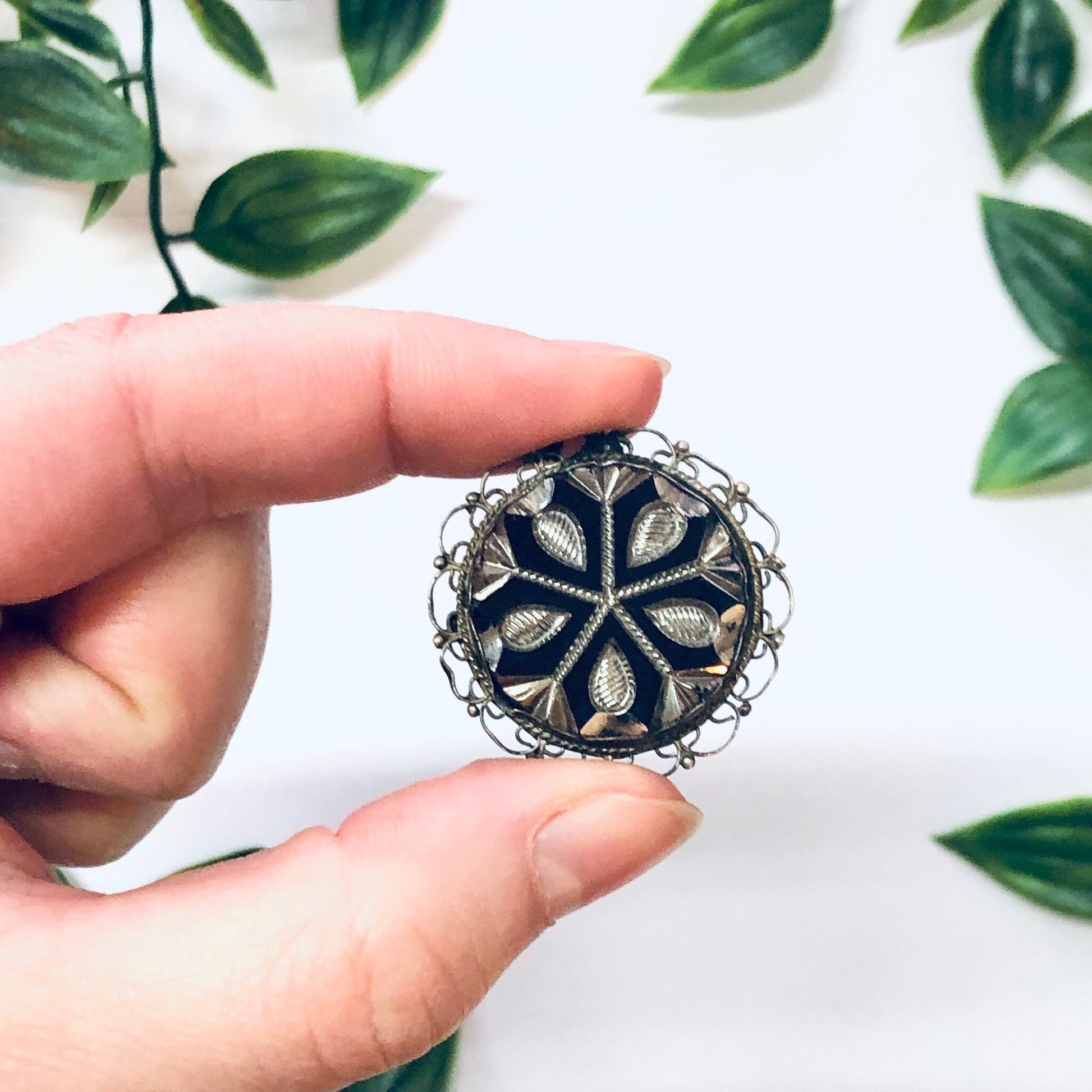 Vintage silver round brooch pendant necklace with unique floral design, held in front of green leaves background