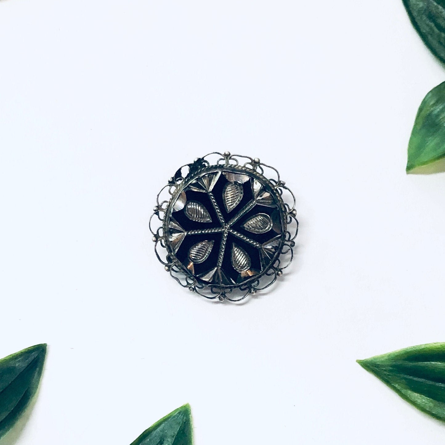 Vintage silver filigree round brooch pendant necklace with intricate cutout design, surrounded by green leaves on white background, unique antique 925 jewelry piece.