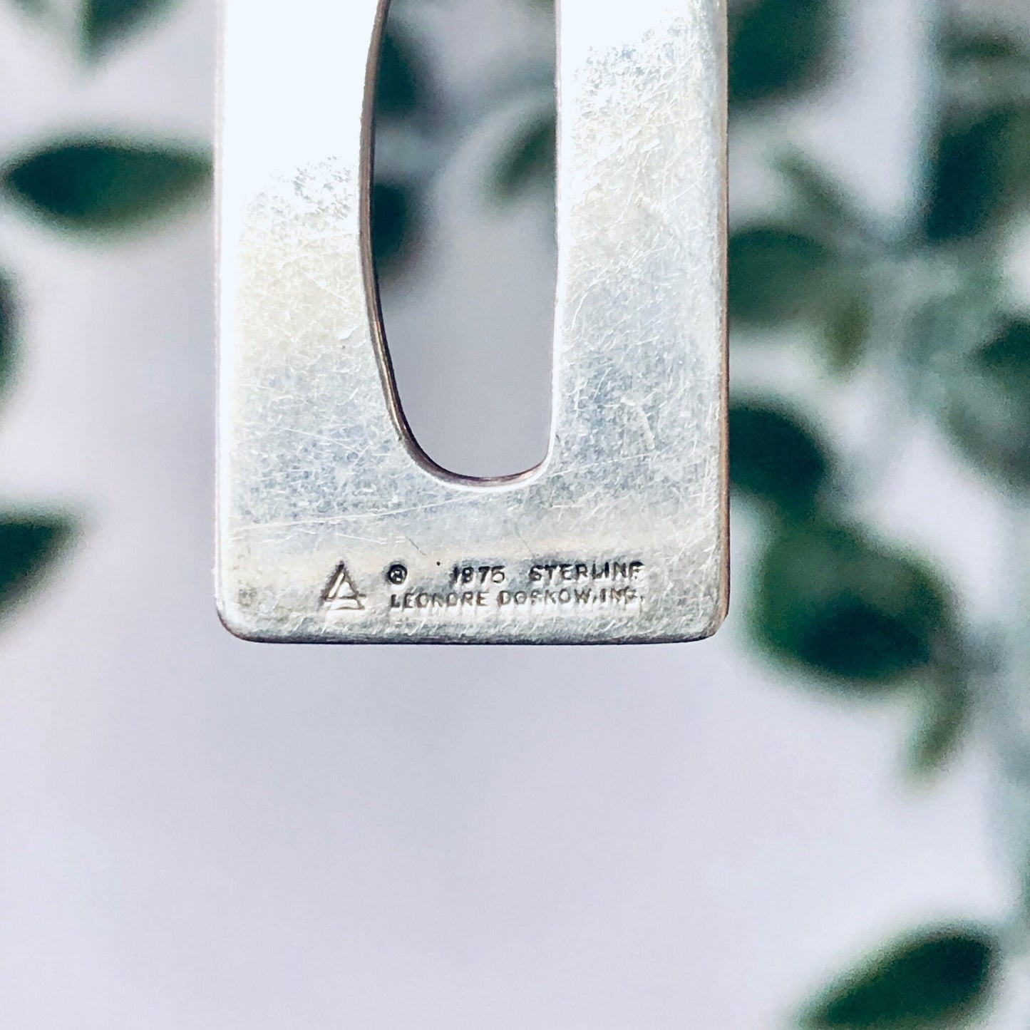 Vintage sterling silver rectangle pendant engraved with "A Hyyro Stfrhinf" and a triangle symbol, photographed against a blurred leafy background.