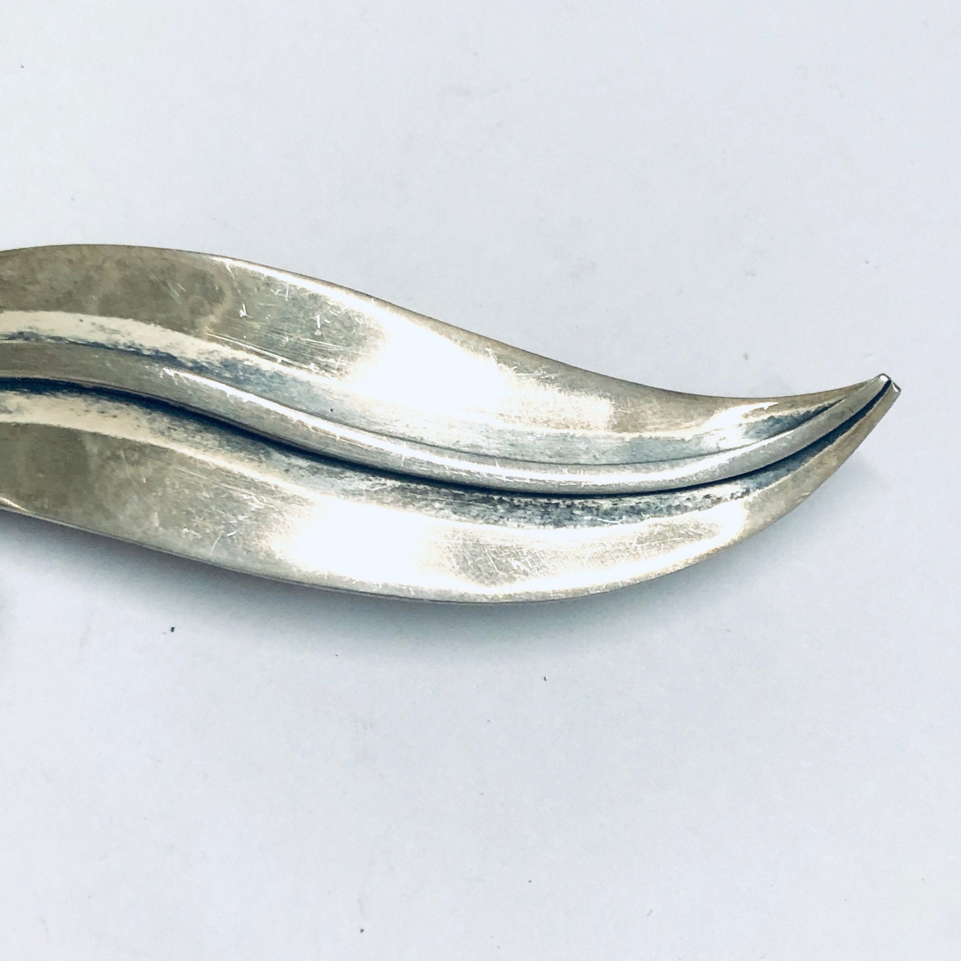 Modernist silver leaf brooch crafted by Delfino in Taxco, Mexico. The large, curved pin resembles a lily of the valley leaf with an organic, flowing shape and textured silver surface that catches the light. An elegant example of midcentury modern Mexican silver jewelry design.