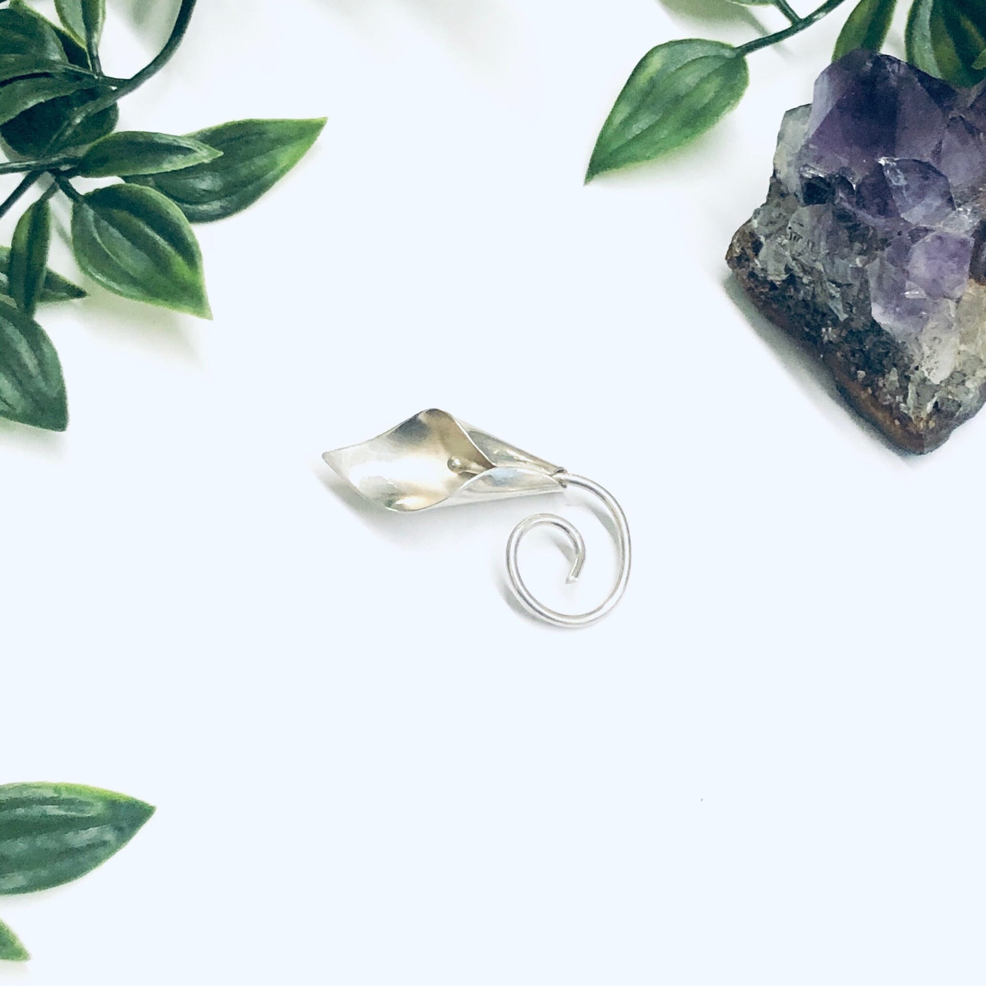 Silver calla lily brooch pin surrounded by green leaves and amethyst crystal on white background