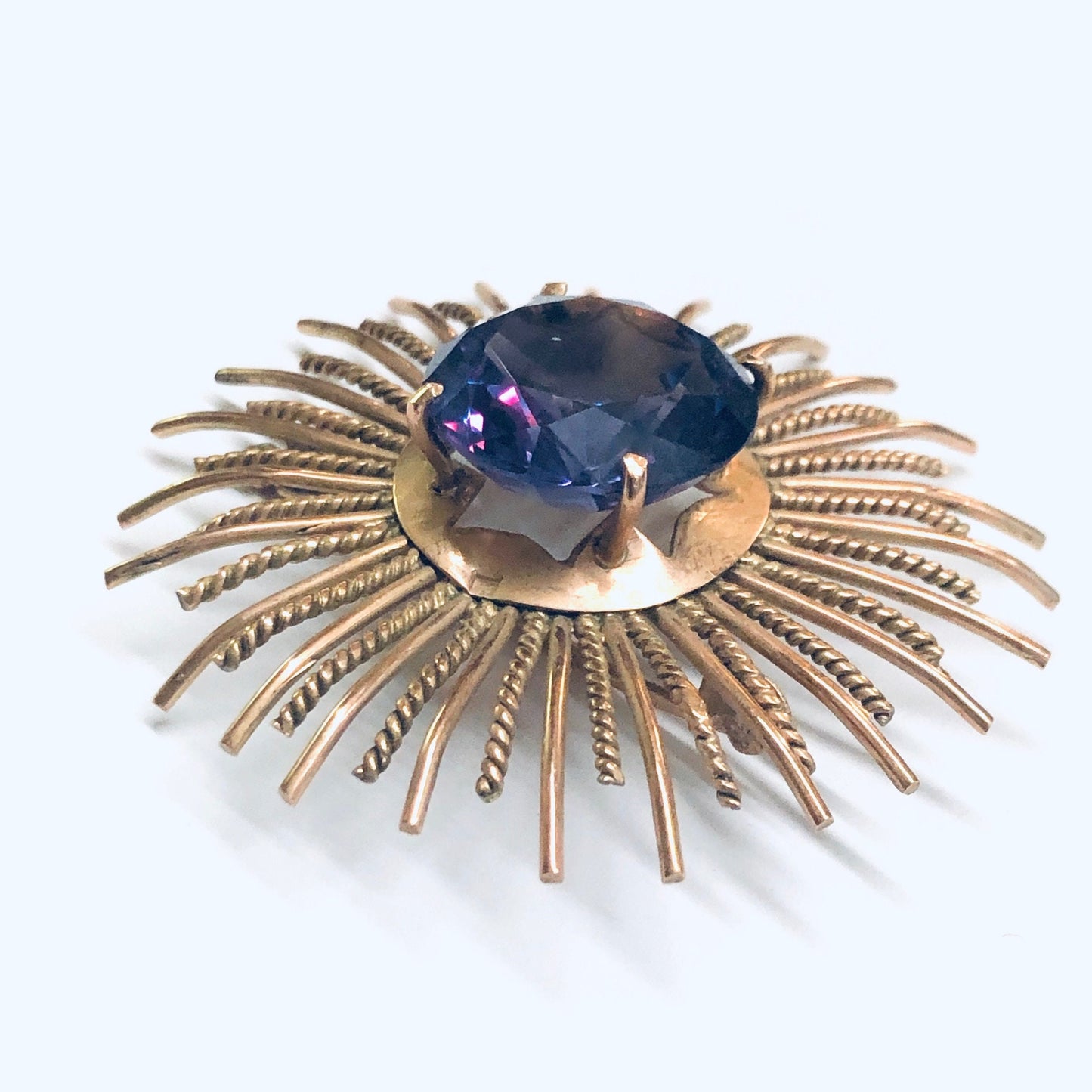 Vintage sunburst brooch in rose gold with a round purple and blue alexandrite gemstone at the center, featuring intricate metal rays radiating outward in a starburst design.