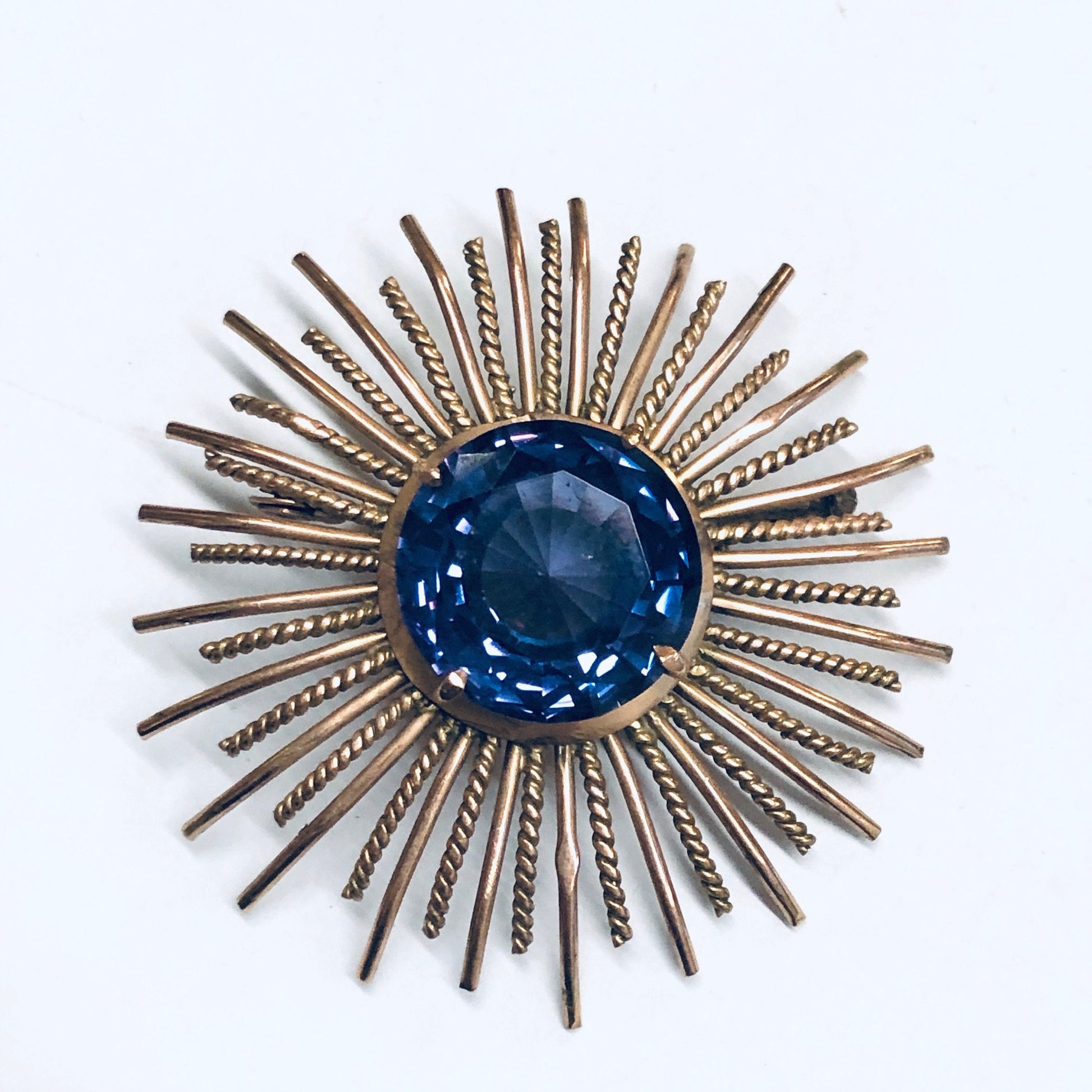 Vintage sunburst brooch with a round blue alexandrite gemstone center, surrounded by radial rose gold bars, creating an elegant purple and blue antique pin or jewelry piece.